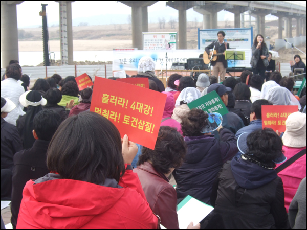 미사 전 공연을 보고 있던 한 참가자가 "흘러라! 4대강! 멈춰라 토건삽질!" 문구가 적힌 플랫카드를 흔들고 있다 / 사진. 평화뉴스 박광일 기자