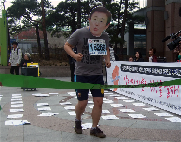 한 장애인단체 회원이 김범일 대구시장의 얼굴이 프린트 된 가면을 쓰고 '장애인 생존권 박탈'이라는 문구가 적힌 결승선을 통과하는 퍼포먼스를 벌이고 있다 / 사진. 평화뉴스 박광일 기자