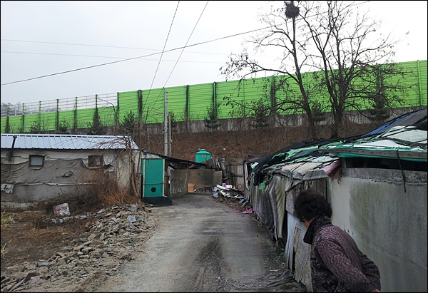 대구시 동구 지저동 886번지 마을. 마을 전체를 감싸고 있는 불투명 소음방지벽과 동구청이 일부 재시공한 투명 소음방지벽. 낮 시간에도 어두운 마을을 바라보는 한조자 할머니(2013.12.30) / 사진.지저동 저지대 주민대책위